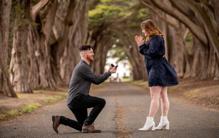 A man proposes to a woman in the middle of a tree lined road.